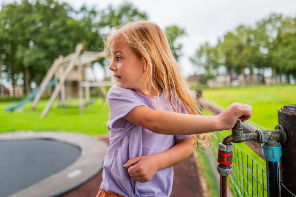 Meisje opent kraan in tuin met trampoline en grasmatten