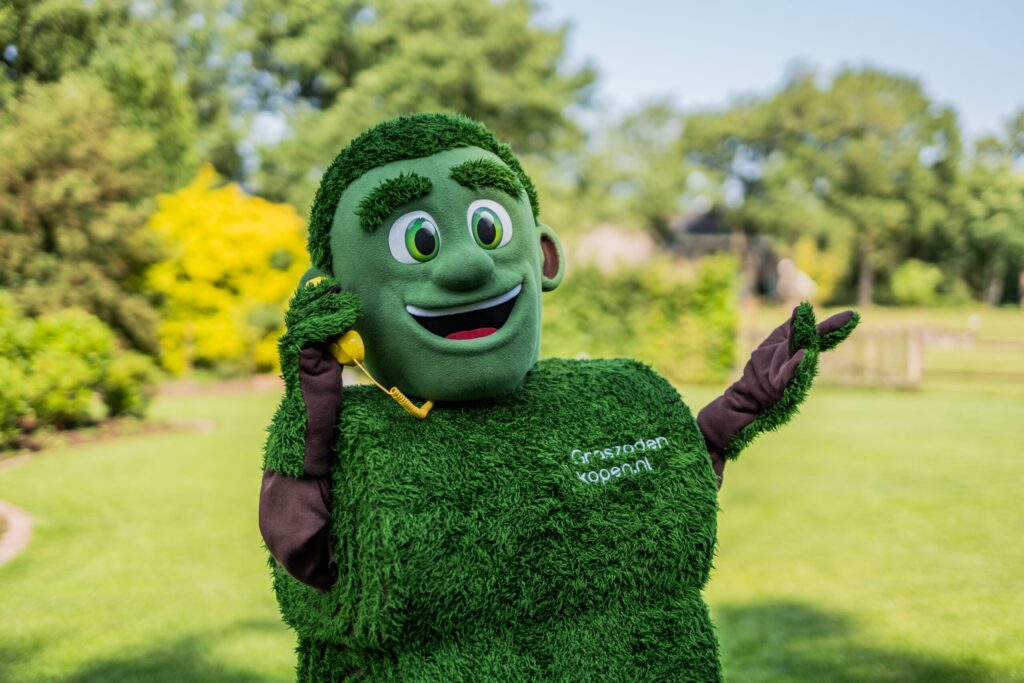 Grasmattie van Graszodenkopen.be belt vrolijk met een gele telefoon in een groene tuin